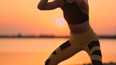 Side-view-of-young-woman-doing-squats-outdoors.-Side-view-of-young-fitness-woman-doing-squats-on-standing-by-the-lake-on-the-sand-at-sunset-the-Slow-motion